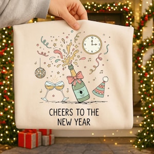May include: A cream-colored tote bag featuring a festive illustration for the New Year. The design includes a champagne bottle with a pink bow, two champagne glasses, a clock, confetti, and the text "CHEERS TO THE NEW YEAR."