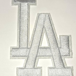May include: A silver embroidered patch featuring the letters "LA" in a stylized design. The letters are filled with a textured, stitched pattern and outlined with a white border. The patch is likely for apparel or accessories.