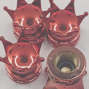 May include: Four red crown-shaped tire valve stem caps. The caps have a shiny, metallic finish and a detailed crown design. One cap is partially disassembled, revealing the internal mechanism.