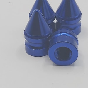 May include: Four blue metal cone-shaped objects with a threaded base. The objects have a glossy finish and are arranged on a white surface. The cones are pointed at the top and have a cylindrical base.
