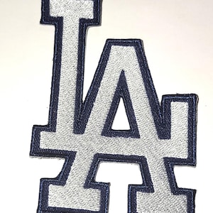 May include: Embroidered patch featuring the letters "LA" in a stylized design. The letters are silver with a navy blue border. The patch is likely for apparel or accessories, representing a team or organization.