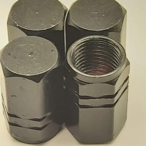 May include: Four black, hexagonal lug nuts. Three are solid, while one has a threaded interior. The lug nuts have a shiny, metallic finish and are designed for automotive use.