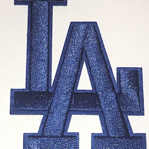 May include: Embroidered patch featuring the letters "LA" in a bold, stylized design. The letters are primarily a vibrant blue with a navy blue border, creating a textured appearance. The patch is likely for apparel or accessories.