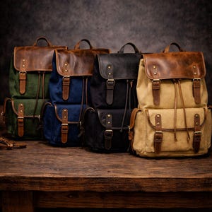 May include: Several canvas and leather backpacks in shades of green, blue, black, and beige. Each bag has a leather top flap, front pockets, and adjustable straps. The backpacks are arranged on a wooden surface.