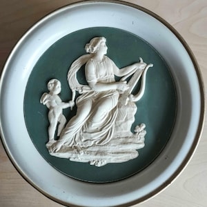 May include: A white ceramic wall plaque with a raised design of a woman playing a lyre and a cherub. The plaque is set against a green background and framed with a white border.