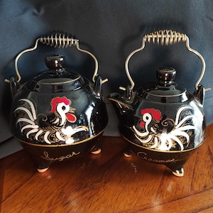 May include: Black ceramic sugar and creamer set with rooster designs. The sugar bowl has the word "Sugar" written on the bottom and the creamer has the word "Creamer" written on the bottom.