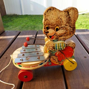 May include: Vintage Fisher-Price pull toy featuring a brown bear playing a xylophone. The bear is wearing a blue and white striped shirt and has a red heart on its chest. The toy has yellow wheels and a red pull string.