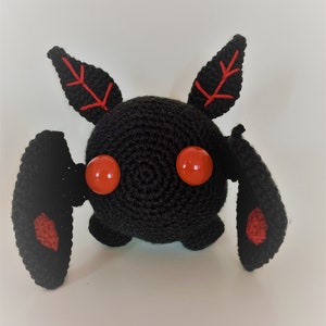 以下が含まれることがあります： 赤い目と耳と羽根に赤いアクセントが付いた黒い毛糸のぬいぐるみ。