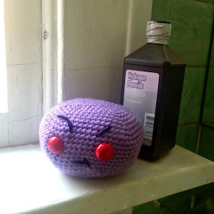 May include: Purple crocheted plush toy with red buttons for eyes. The toy is sitting on a white surface next to a bottle of hydrogen peroxide.