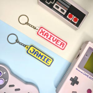 May include: Two pixelated keychains with the names "Xaiver" and "Jamie" in white letters. The "Xaiver" keychain is red, and the "Jamie" keychain is yellow. The keychains are on a chain with a keyring. The background includes retro video game controllers.
