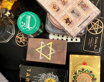 May include: A collection of tarot decks, including a deck with a gold and green mandala design, a deck with a black background and a gold eye design, and a deck with a brown background and a gold star of David design. The decks are on a black surface with other items, including a glass jar, a small green container, and a black figurine.