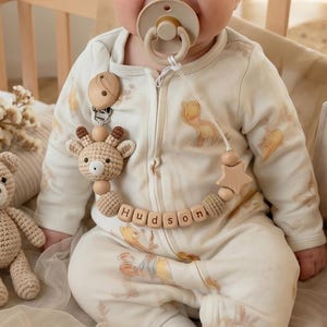 May include: A baby in a wooden crib wearing a beige onesie with a giraffe and animal print. The baby has a pacifier and a wooden pacifier clip with the name "Hudson". A crocheted teddy bear and books are also in the crib.