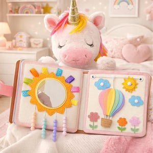 May include: A plush unicorn with a golden horn rests above an open, soft activity book. The book features a mirror, sun, and colorful ribbons on one page, and a hot air balloon, sun, and flowers on the other. The book is designed for babies and toddlers.