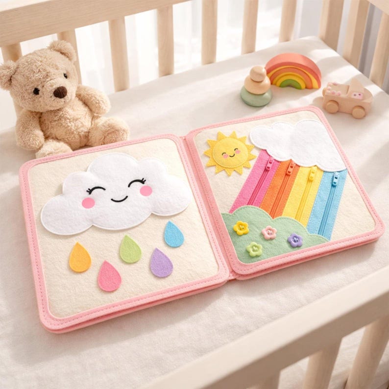 May include: A soft, open book with felt illustrations of a smiling cloud with raindrops and a sun with a rainbow. The book is pink-edged and sits on a white surface, with a teddy bear and wooden toys nearby.