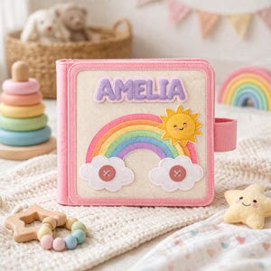 May include: A pink felt activity book with the name "AMELIA" on the cover. The cover features a rainbow, smiling sun, and clouds. Other toys include a wooden star teether, a star-shaped plush, and a rainbow-colored stacking toy.