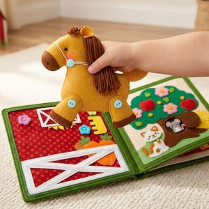 May include: A felt farm-themed activity book with a plush horse toy. The book features a red barn scene with a white gate and a green-bordered page with a tree, flowers, and a cat. The horse is tan with a brown mane and blue button accents.