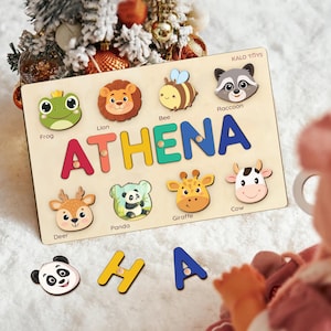 May include: Wooden puzzle board with the name "ATHENA" spelled out in colorful letters. The board features animal-shaped puzzle pieces including a frog, lion, bee, raccoon, deer, panda, giraffe, and cow. The brand name "KALO TOYS" is visible.