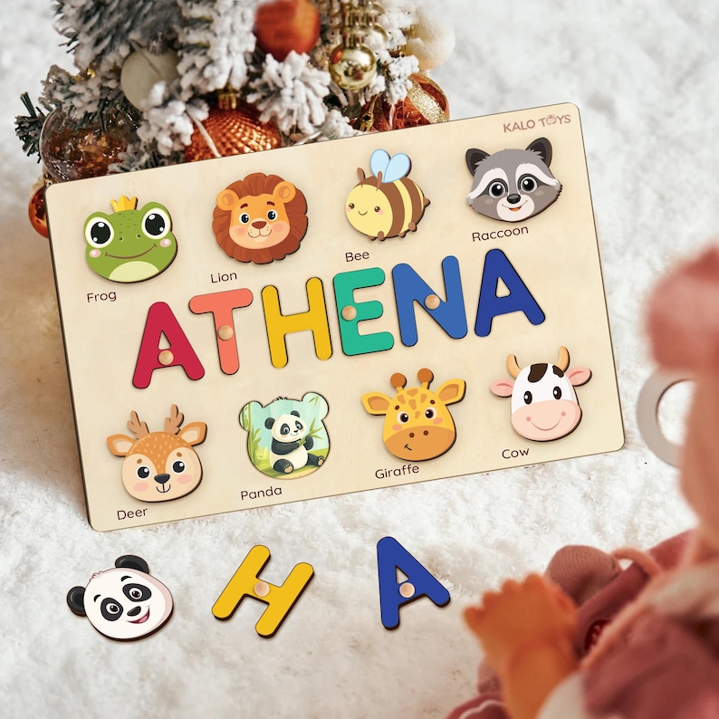 May include: Wooden puzzle board with the name "ATHENA" spelled out in colorful letters. The board features animal-shaped puzzle pieces including a frog, lion, bee, raccoon, deer, panda, giraffe, and cow. The brand name "KALO TOYS" is visible.