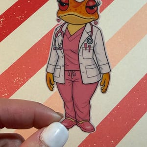 Puede incluir: Una pegatina con una rana de dibujos animados vestida con ropa médica. La rana lleva un uniforme rosa, una bata blanca y un estetoscopio. La rana tiene la piel naranja, pestañas rojas y pendientes en forma de corazón.