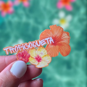 Op de afbeelding: Een sticker met het woord "TROPICOQUETA" in het wit, vergezeld van een tropisch bloemdesign. Het ontwerp bevat een oranje hibiscusbloem, een rode bloem en een gele bloem. De achtergrond is een wazige turquoise kleur.