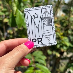 May include: A white and black sticker with a Puerto Rican flag design and the letters "PR". The sticker features a star, a building, and a flag design. The sticker is held in front of a green leafy background.