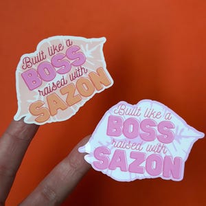 May include: Two stickers with the text "Built like a BOSS raised with SAZON" in pink and orange. The stickers are in the shape of a stylized mouth or lips, with a white outline. The background is orange.