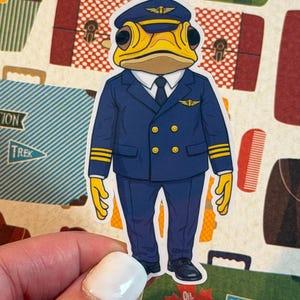Puede incluir: Una pegatina con una rana de dibujos animados vestida con un uniforme de piloto azul con detalles dorados. La rana lleva un gorro de piloto y una corbata. La pegatina está sujeta por una mano, con fondos temáticos de viajes.