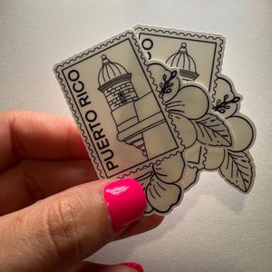 May include: Two stickers featuring a black and white design of a Puerto Rican landmark and a flower. The stickers are in the shape of postage stamps, with the words "Puerto Rico" printed vertically on one. The stickers are held in a hand.