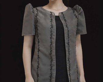 May include: A grey open-front jacket with short puffed sleeves and black floral embroidery along the vertical edges, worn over a simple black dress. The jacket features a round neckline and a structured silhouette with a smooth, semi-sheer fabric finish.