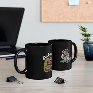 Può includere: Due tazze in ceramica nera con grafiche a cartoni animati. Una tazza presenta la parola "ALPHA" con un personaggio dei cartoni animati. L'altra tazza ha un'immagine di cartone animato e il testo "Money Over Everything".