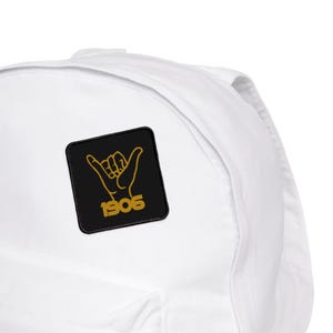 May include: A white backpack with a black square patch. The patch features a gold hand gesture and the number "1906" in gold. The backpack is made of a smooth, durable material.