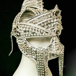 May include: A silver-colored, ornate helmet with a spiked crest and intricate detailing. The helmet is covered in small, reflective, geometric shapes, and features decorative elements. The helmet is displayed on a white mannequin head against a black background.