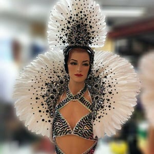 May include: White feather headdress with black polka dots and rhinestones. The headdress is worn by a mannequin with a white feather boa and a sequined costume.