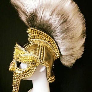 May include: A gold-colored gladiator helmet with a spiked design and a long, white and gray feather plume. The helmet is decorated with gold beads and is on a white mannequin head.