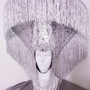 May include: A silver sequined and beaded headdress with a wide brim and long fringe. The headdress is worn on a white mannequin with a silver beaded necklace.