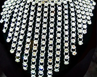 May include: A striking silver necklace with a bib design. The necklace is adorned with numerous clear, sparkling crystals arranged in a chain-like pattern. The crystals cascade downwards, creating a glamorous effect. The background is black, enhancing the necklace's sparkle.