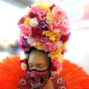 May include: A mannequin head adorned with a large, colorful floral headdress. The face is covered by a jeweled skull mask. The mannequin is wearing an orange feathered collar and embellished shoulder pieces, creating a vibrant and theatrical look.