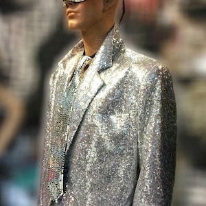Puede incluir: Un maniquí con una americana plateada con lentejuelas, corbata a juego y una máscara de espejo. La chaqueta tiene un corte clásico con solapas con muescas. La máscara cubre los ojos y la nariz. El look general es glamuroso y teatral.