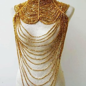 Puede incluir: Un collar de cuentas doradas con diseño de gargantilla de cuello alto, compuesto por múltiples hileras de pequeñas cuentas doradas reflectantes que caen en cascada sobre el pecho en un patrón semicircular superpuesto, creando una pieza de joyería.
