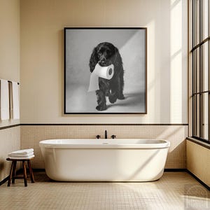 Puede incluir: Impresión en blanco y negro de un perro negro sosteniendo un rollo de papel higiénico. El perro es el foco central, sobre un fondo neutro. La impresión cuelga sobre una bañera blanca en un baño.