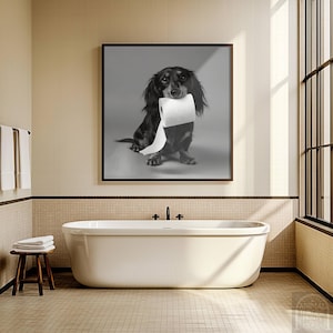 May include: Black and brown dachshund dog holding a roll of toilet paper in its mouth, framed in a black and white photograph. The artwork is displayed above a white bathtub in a bathroom setting.