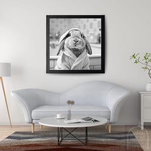 May include: Black and white framed print of a rabbit wearing a hooded robe. The rabbit is centered in the frame, with its ears and face visible. The background is a blurred bathroom scene. The print is displayed above a gray sofa.