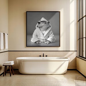 May include: Black and white framed print of a frog wearing a white bathrobe and hood. The frog is sitting with its arms crossed. The print is hanging above a white bathtub in a bathroom setting.