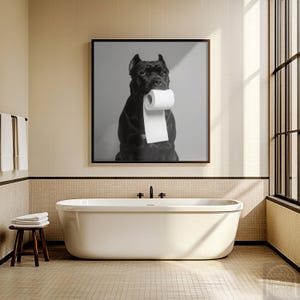 May include: A black dog with a roll of toilet paper in its mouth, in a black and white photo, hanging on a wall above a white bathtub in a bathroom.