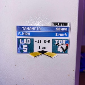 Può includere: Un magnete a tema baseball con i nomi YAMAMOTO e KIRK e la parola SPLITTER. Il magnete mostra i punteggi di LAD e TOR, con la scritta "1 OUT". Il magnete è blu, bianco e oro.