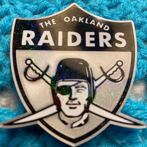 May include: A shield-shaped Oakland Raiders emblem with the words "THE OAKLAND RAIDERS" in white lettering. The emblem features a silver and black color scheme with a football player's helmet and crossed swords.
