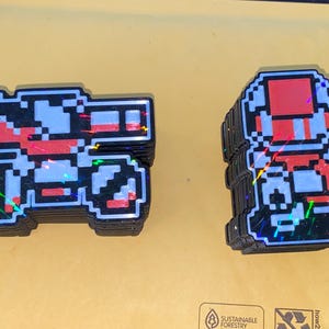 Könnte beinhalten: Stapel von Pixel-Art-Aufklebern mit einer roten und blauen Videospiel-Figur und einem Fahrzeug. Die Aufkleber haben ein holografisches, irisierendes Finish und befinden sich auf einem gelben Hintergrund. Die Designs erinnern an Retro-Videospiele.