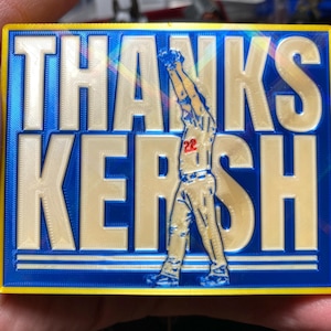 May include: A rectangular blue and yellow plaque with the words "THANKS KERSH" in large, raised, off-white letters. A baseball player in a baseball uniform is depicted in the center. The player is wearing the number 22.