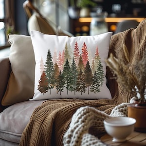 May include: White square throw pillow featuring a watercolor illustration of a forest of pine trees in shades of green, brown, and red. The pillow is placed on a sofa with a brown blanket.