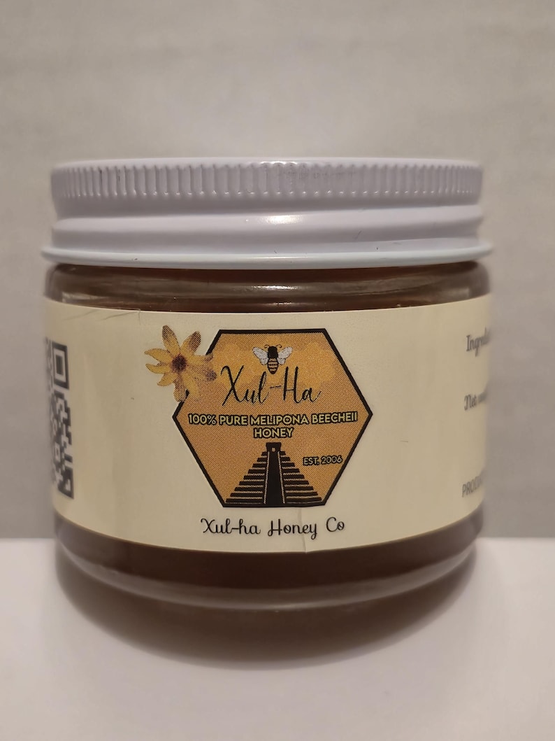 K&ouml;nnte beinhalten: Ein Glas Honig mit einem wei&szlig;en Deckel. Das Etikett ist gelb und zeigt eine Biene, eine Pyramide und den Text "Xul-Ha 100% Pure Melipona BeeCheii Honey Est. 2006 Xul-ha Honey Co."